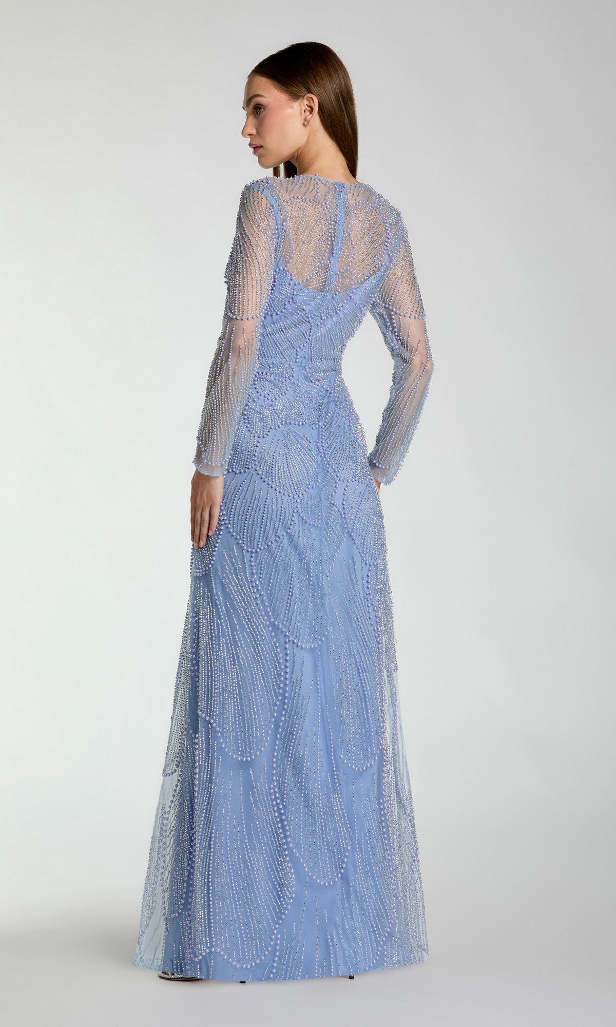A woman faces away, wearing the Mac Duggal 69012—a floor-length, long-sleeved, light blue modest formal dress adorned with intricate beaded embellishments.